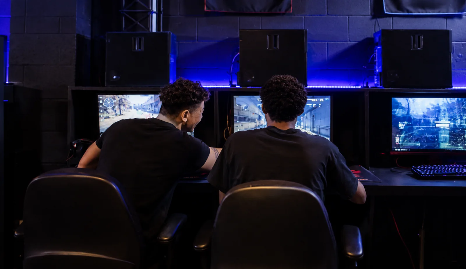 Two gamers seated at computer stations in a dimly lit gaming room, playing competitive shooter games under blue LED lighting.