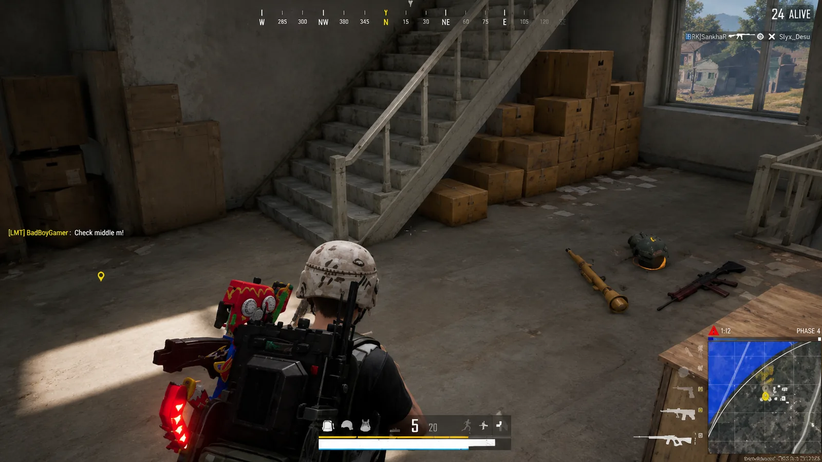 PUBG gameplay showing an armed player inside a building near a staircase, with weapons, equipment, and loot scattered across the floor.
