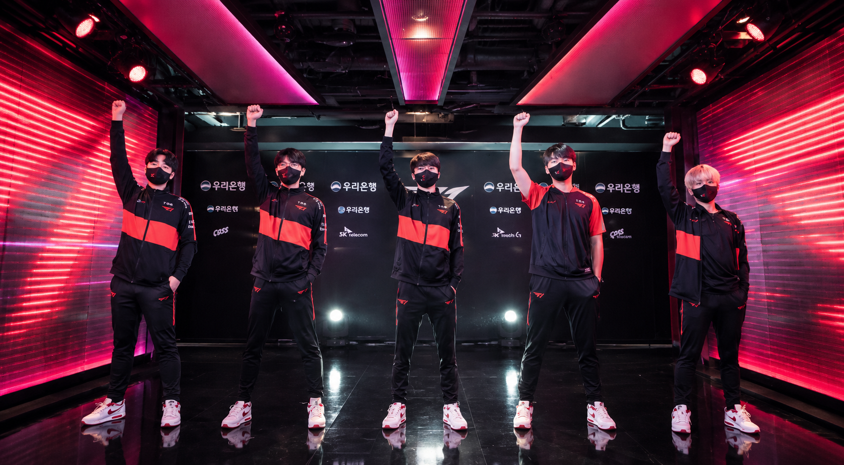 Five masked South Korean esports players in matching black-and-red team outfits raise their fists in a futuristic arena corridor during an esports event.
