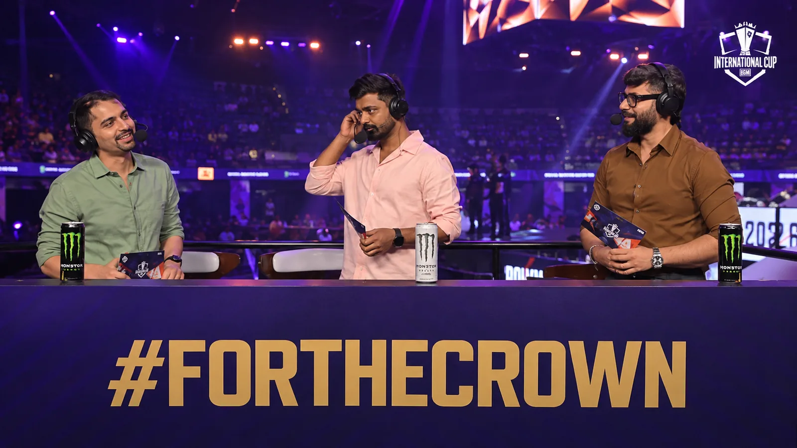 Three esports commentators present the BMIC 2026 broadcast from the tournament arena, with the #FORTHECROWN slogan displayed across the desk.